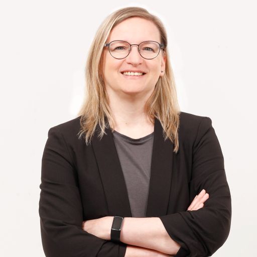 Frau mit Brille und blonden Haaren, schwarzem Blazer, verschränkten Armen, lächelnd vor weißem Hintergrund.