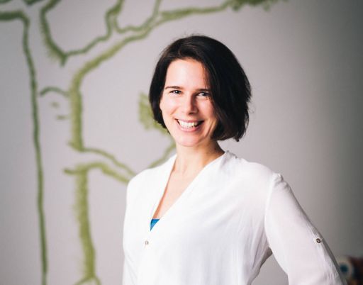 Frau mit kurzem dunklem Haar, weißer Bluse und blauem Top lächelt vor einer Wand mit grünem Baumgemälde.