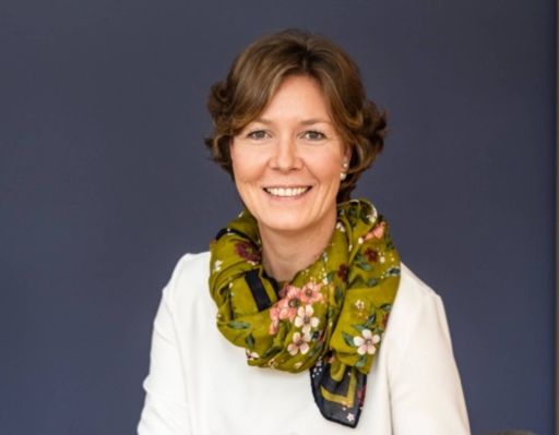 Frau mit kurzen braunen Haaren, weißem Blazer und grün-geblümtem Schal lächelt vor grauem Hintergrund.