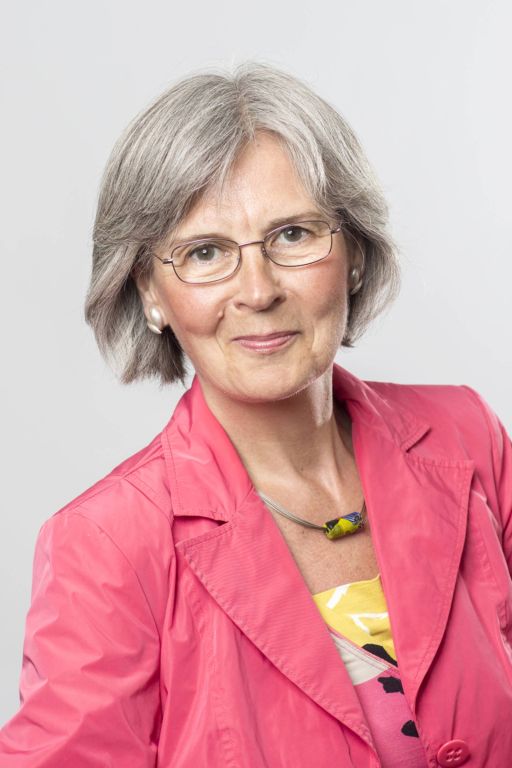 Ältere Frau mit grauem Bob, Brille und rosa Blazer lächelt vor hellem Hintergrund in die Kamera.