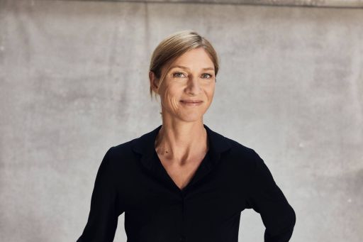 Frau mittleren Alters mit blondem Haar, schwarzer Bluse, lächelnd vor grauer Betonwand stehend.