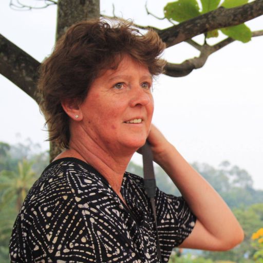 Ältere Frau mit kurzen braunen Haaren in gemusterter Bluse steht unter einem Baum, Blick zur Seite.