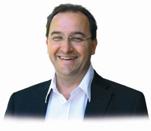 Mann mit Brille und dunklem Sakko über weißem Hemd lächelt breit vor weißem Hintergrund.