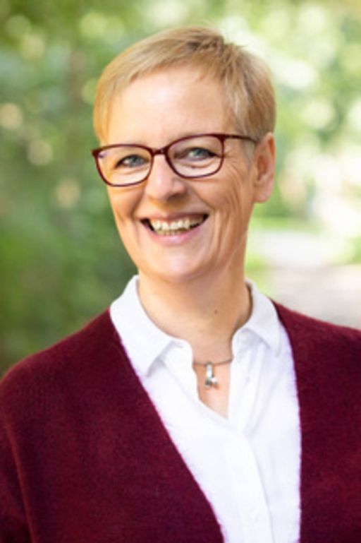 Lächelnde Frau mit kurzen blonden Haaren und roter Brille, trägt bordeauxrote Strickjacke, vor grünem Hintergrund.