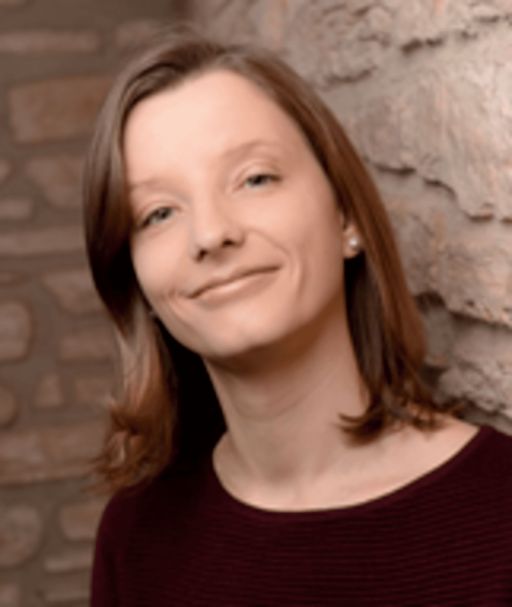 Frau mit schulterlangem braunem Haar und burgunderrotem Pullover lächelt vor einer Steinmauer.