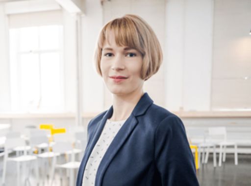Frau mit kurzem blondem Bob und blauem Blazer steht lächelnd in einem hellen, modernen Raum mit weißen Stühlen.