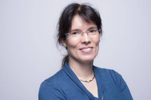 Frau mit dunklem Haar, Brille und blauem Oberteil lächelt vor hellem Hintergrund.