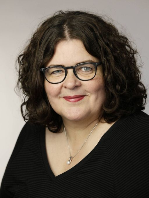 Frau mit dunklen Locken und schwarzer Brille, schwarzem V-Ausschnitt-Oberteil und silberner Halskette, lächelt.