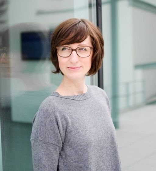 Frau mit kurzem braunem Haar und Brille in grauem Pullover steht vor einer Glasfassade.