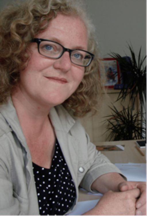 Frau mit lockigem, blondem Haar und schwarzer Brille sitzt an einem Schreibtisch, trägt ein gepunktetes Oberteil.