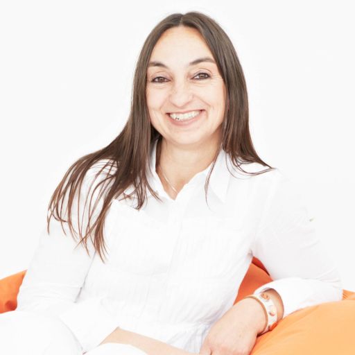 Frau mit langen braunen Haaren, weißem Hemd und Armband sitzt lächelnd auf orangefarbenem Sitzsack.