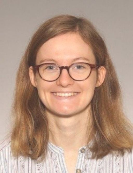 Porträtfoto einer Person mit schulterlangem rötlich-braunem Haar und runder Brille, lächelnd, helles Oberteil.