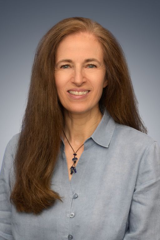 Frau mit langen braunen Haaren, hellblauem Hemd und Steinkette lächelt vor grauem Hintergrund.