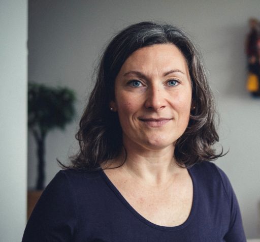 Frau mit schulterlangem dunkelbraunem Haar und blauen Augen, trägt dunkelblaues T-Shirt, lächelt leicht, Innenraum.