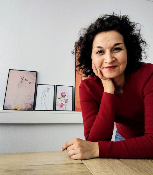 Frau mit schwarzen Locken im roten Pullover sitzt an Holztisch, dahinter Regal mit drei gerahmten Kunstdrucken.