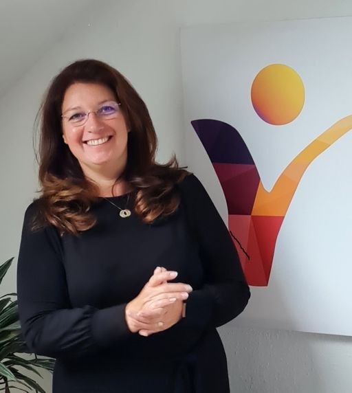 Lächelnde Frau mit Brille und dunklem Oberteil steht vor einem bunten Logo mit menschlicher Figur.