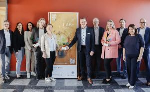 Gruppe von ca. 11 Personen steht vor einem Banner mit Aufschrift „REGIO LAB+" in einem Gebäude.