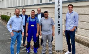 Sechs Männer stehen vor dem Schild „Haus der Wirtschaftsförderung". Zwei tragen Arbeitskleidung, die anderen Freizeitkleidung.
