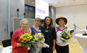 Vier Frauen stehen nebeneinander in einem Konferenzraum, drei halten Blumensträuße, im Hintergrund eine Leinwand.