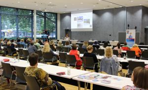 Konferenzraum mit vielen Personen an Tischen, eine Rednerin vor Leinwand mit Präsentation, große Fensterfront.