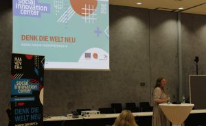 Frau spricht an einem Stehtisch vor einer Leinwand mit „Denk die Welt Neu"-Präsentation des Social Innovation Centers Hannover.