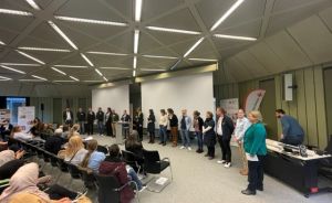Gruppe von Personen steht in einem Konferenzraum vor Publikum; im Hintergrund ein Rotes-Kreuz-Banner.