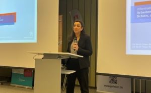 Frau am Rednerpult mit Mikrofon, zwei Projektionsleinwände mit „Hannover"-Logo, Veranstaltungsraum.