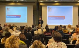 Person hält Vortrag vor vollem Publikum in Konferenzraum. Zwei Projektionsflächen zeigen „Hannover"-Präsentation.