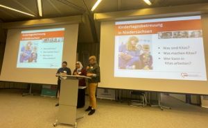 Drei Personen stehen an einem Rednerpult vor zwei Projektionsflächen mit deutscher Präsentation über „Kindertagesbetreuung in Niedersachsen".