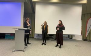 Drei Frauen stehen in einem Konferenzraum; eine hält ein Mikrofon, links ein Rednerpult, hinten eine Leinwand.