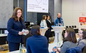 Frau in blauem Blazer spricht mit drei sitzenden Frauen bei einer Netzwerkveranstaltung in einem Konferenzraum.