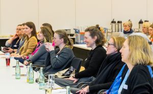 Mehrere Frauen sitzen in einer Reihe an einem langen Tisch in einem Konferenzraum und hören aufmerksam zu.