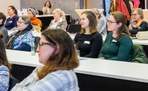 Mehrere Frauen sitzen in Reihen an weißen Tischen in einem Konferenzraum und schauen aufmerksam nach vorne.