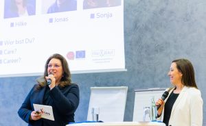 Zwei Frauen stehen an Stehtischen mit Mikrofonen vor einer Präsentationsfolie mit Namen und Logos.