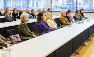 Gruppe von Personen sitzt in Reihen an langen weißen Tischen in einem Konferenzraum und hört aufmerksam zu.
