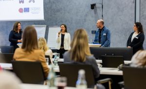 Vier Personen stehen vor Publikum in einem Konferenzraum. Eine Frau spricht ins Mikrofon, hinter ihr eine Präsentation.