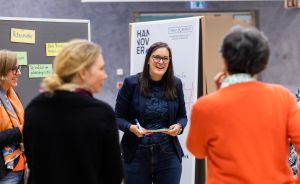 Frau in blauem Blazer hält Stift und Buch, umgeben von drei Personen vor Pinnwand mit "Hannover"-Banner.