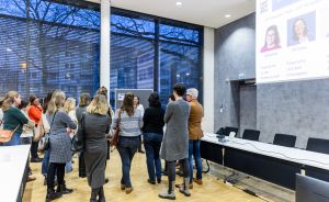 Gruppe von Personen steht in einem modernen Konferenzraum vor einer Präsentation. An der Wand ein Bildschirm mit Porträts.