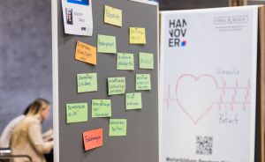Pinnwand mit bunten Haftnotizen auf Deutsch, daneben ein Plakat mit „HANNOVER" und einem Herz mit „Familie & Beruf".