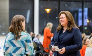 Zwei Frauen im Gespräch bei einer Veranstaltung; eine hält ein Mikrofon, im Hintergrund weitere Personen.