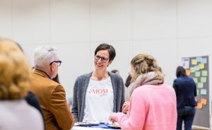 Drei Personen im Gespräch an einem Stehtisch. Frau in der Mitte trägt weißes T-Shirt mit Aufdruck „#MOM".