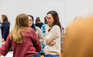 Drei Frauen stehen in einem hellen Raum und unterhalten sich. Eine trägt eine Brille und einen beigen Pullover.