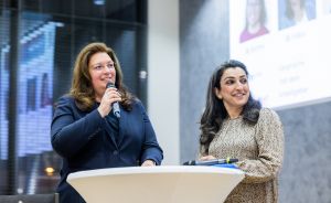 Zwei Frauen stehen an einem Stehtisch, eine spricht ins Mikrofon, im Hintergrund eine Präsentationsleinwand.