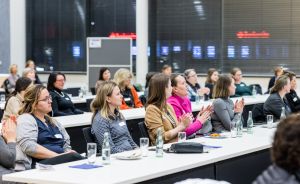 Frauen sitzen an langen Tischen in einem Konferenzraum und klatschen. Wassergläser und Notizen auf den Tischen.