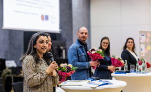 Frau spricht ins Mikrofon an Rednerpult. Im Hintergrund stehen drei Personen mit Blumensträußen vor Leinwand.