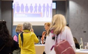 Frauen stehen in einem Konferenzraum und unterhalten sich. Im Hintergrund eine Leinwand mit Silhouetten.