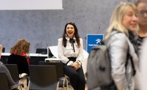 Frau in weißer Bluse und schwarzer Hose sitzt lachend auf einem Stuhl in einem Konferenzsaal.