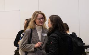 Zwei Frauen mit Namensschildern unterhalten sich auf einer Veranstaltung; im Hintergrund weitere Personen.