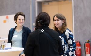 Drei Frauen stehen an einem Stehtisch in einem Konferenzraum und unterhalten sich. Rechts ein roter Feuerlöscher.