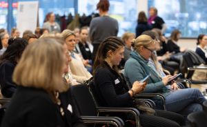 Publikum sitzt in Stuhlreihen bei einer Veranstaltung; mehrere Frauen schauen auf Smartphones und Tablets.